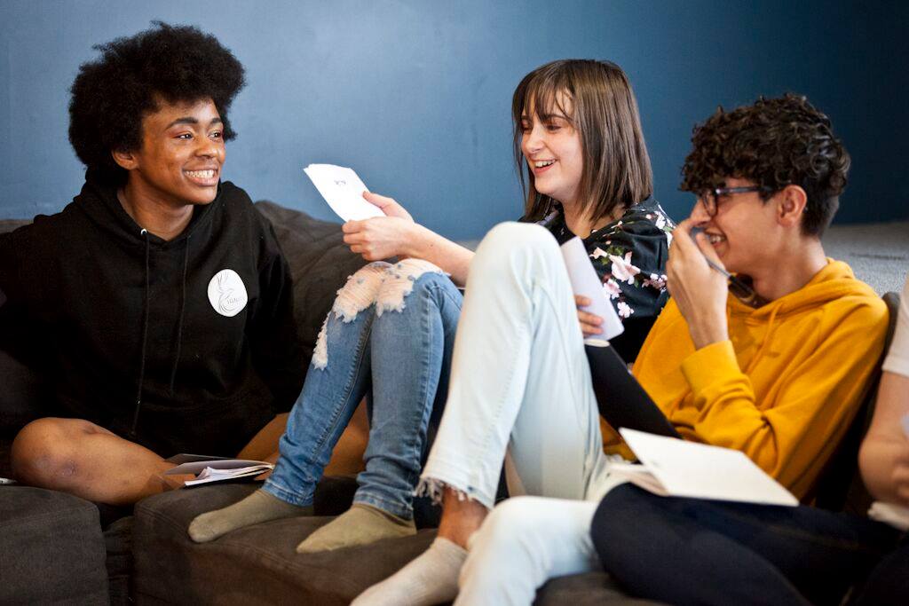 Three young people sitting together, holding pieces of paper and laughing. Ignite Theatre.