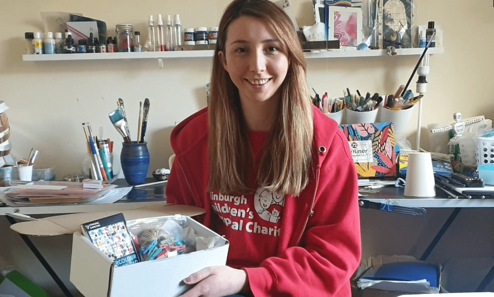 A red-haired young person smiling at the camera holds an Edinburgh Children's Hospital Charity arts and crafts studio box.