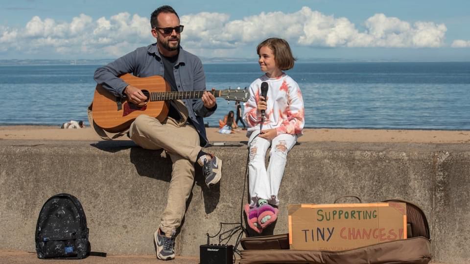 A man sits on a wall playing the guitar, a 9 year old girl child is holding a microphone and smiling.