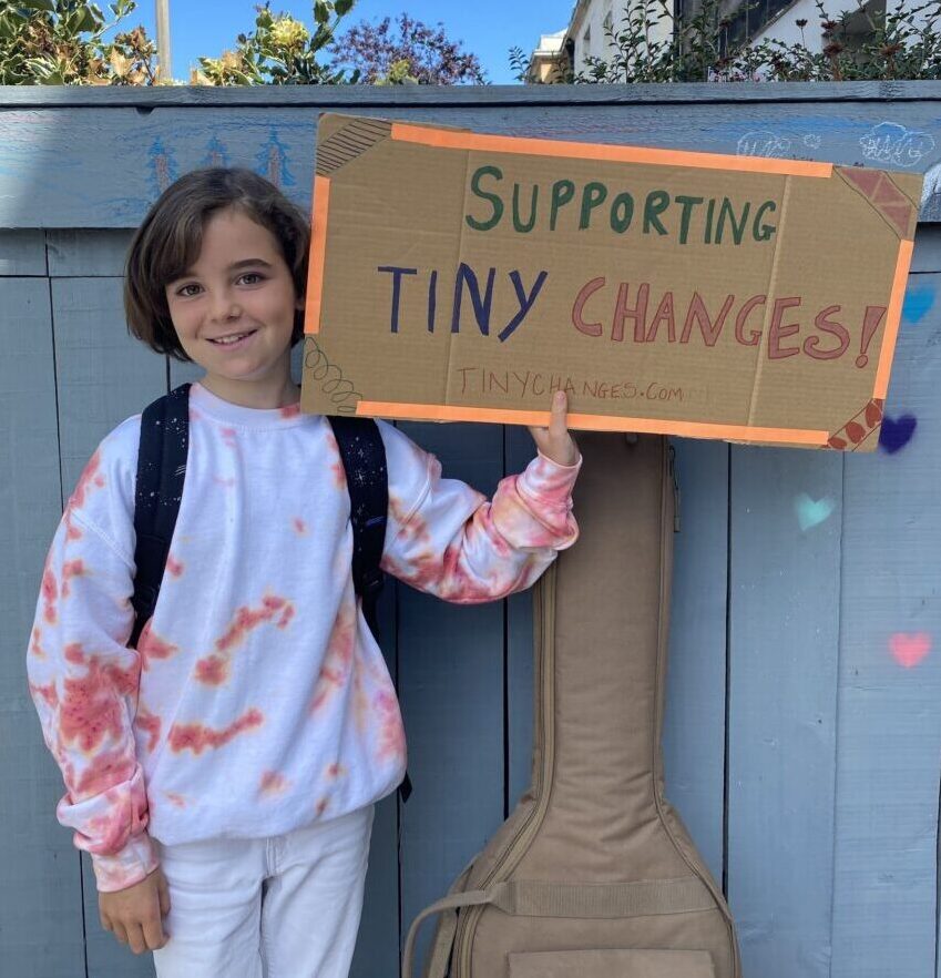 A young girl child smiles at the camera holding a card board sign that says 'Supporting Tiny Changes'. 