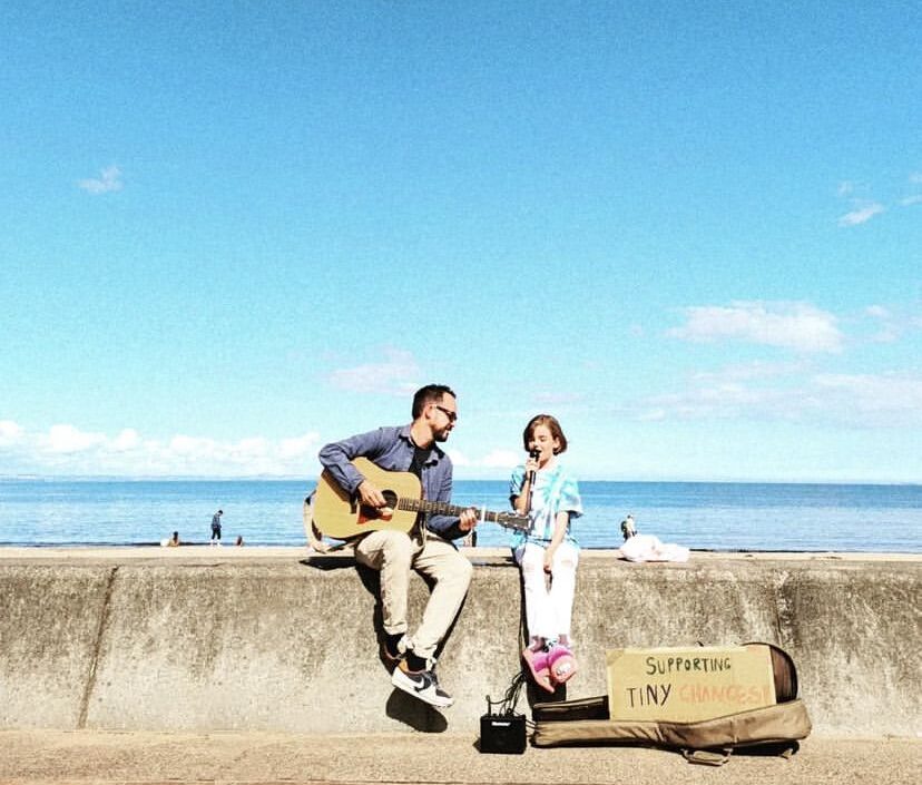 A man sits on a wall playing the guitar looking at a young girl child who is holding a microphone.